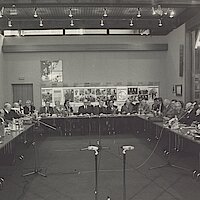 Beteiligte der Konferenz sitzen an Tischen, zwischen denen zahlreiche Mikrofone aufgebaut sind.