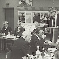 Ein Mann spricht an einem Rednerpult, um ihn herum sitzen an einem Konferenztisch weitere Männer
